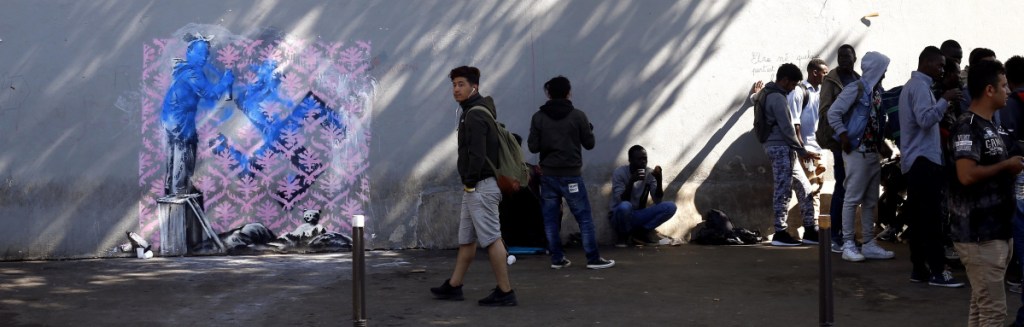 Graffiti attributed to street artist Banksy appears on a wall near a group of Paris migrants. It depicts a child, standing on an object at left, spray-painting wallpaper over a swastika.