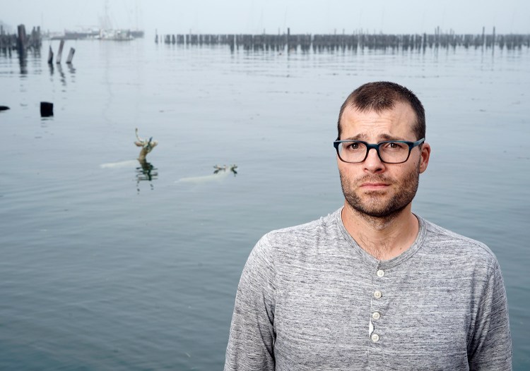 Two deer sculptures that Andy Rosen placed in Portland Harbor near the Ocean Gateway terminal in Portland are visible behind him. His project, funded with a grant from the Union of Concerned Scientists, illustrates that rising sea levels are claiming land that animals and people depend on.