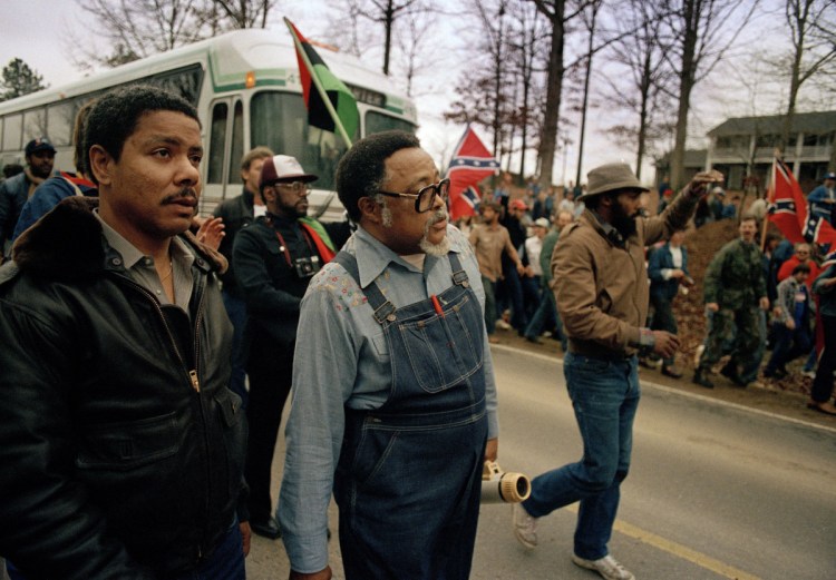 In this Jan. 18, 1987, photo, Atlanta city councilman, Rev. Hosea Williams, in overalls, leads a march against efforts to keep Forsyth County in Georgia all white past counter-protesters near Cumming, Ga., as a crowd waves Confederate flags and jeer the marchers. Racial stereotypes and racist imagery in popular culture seemed to be everywhere in the chaotic 1980s when future Virginia Gov. Ralph Northam and future Attorney General Mark Herring admitted dressing up in blackface. 