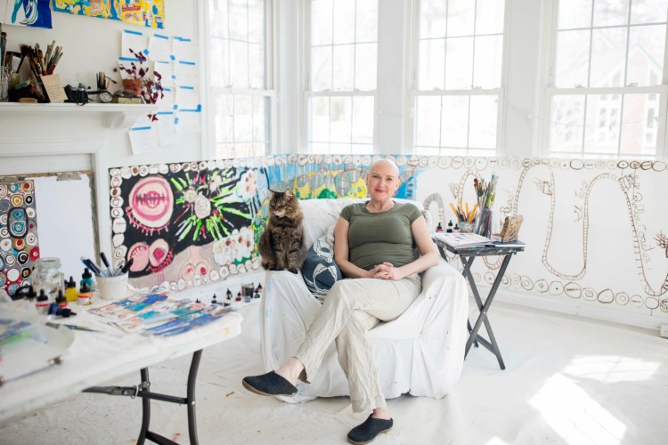 Erin McGee Ferrell, with her cat, Milo, in her studio at home in Falmouth. McGee Ferrell was diagnosed with breast cancer and is currently going through chemotherapy treatments. She has created a series of paper dolls with names like Ms. Mastectomy and Ms. Radiation as an educational series to help women navigate their cancer treatments.