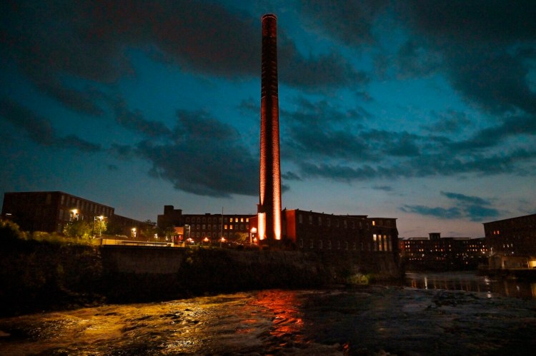 The Pepperell Mill Campus' smokestack is lit up at night along the Saco River in Biddeford in October. Two local developers are purchasing the property as an investment.