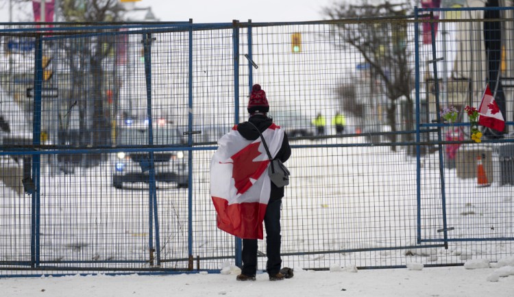 A lone protester stands draped in the Canadian flag at a temporary fence controlling access to streets near Parliament Sunday in Ottawa.