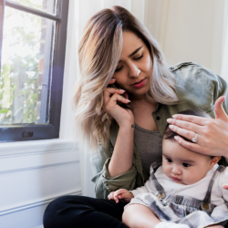 A woman holds one hand to her baby's forehead while she holds a phone to her ear.