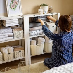 A woman in a blue shirt sits in front of open shelves, organizing linens and bathroom objects into smaller baskets.