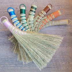 Seven broomcorn whisks with multi-colored handles fan out on a wood background.