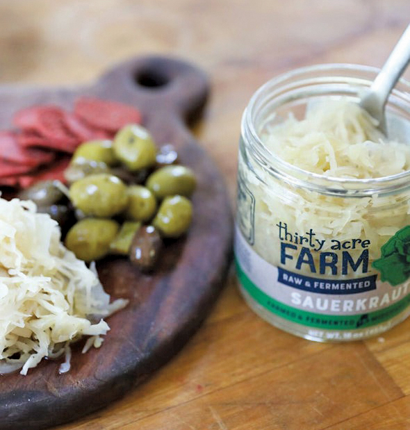 An open container of Thirty Acre Farm sauerkraut next to a charcuterie board that includes a scoop of kraut.